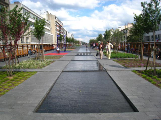 Aménagement urbain aquatique agrémenté de vegetation ou se mêle des allées pavées de dalles noires.